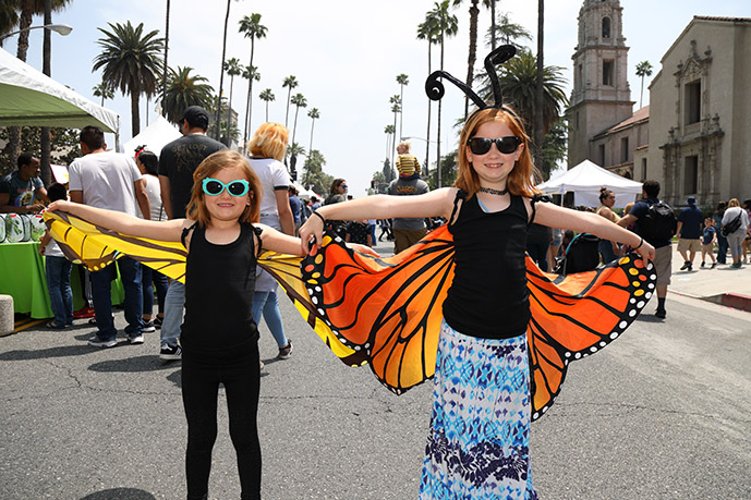 A photo of two girls both smiling and standing in front of a booth showcasing their butterfly wing costume inspired by Pink Lady Butterflies at the Riverside Insect Fair.  